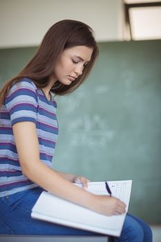 Portrait of schoolgirl sitting on bench and writing on book in classroom at school