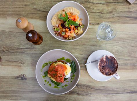 Delicious breakfast plates of smoked salmon poached eggs on toast and fresh fruit salad with yoghurt and cappuccino.   Gourmet food, flavour, tasty, mouthwatering, hungry, flatlay