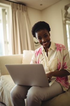 Portrait of smiling woman sitting on sofa and using laptop in living room at home