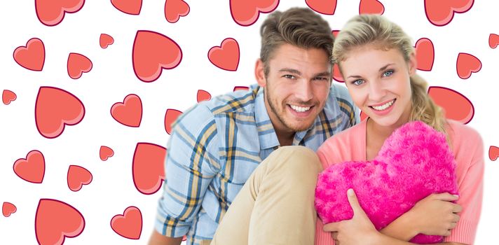 Attractive young couple sitting holding heart cushion against background with hearts