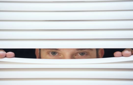 Caucasian man looking through the slats of a blind themes of curiosity watching surveillance