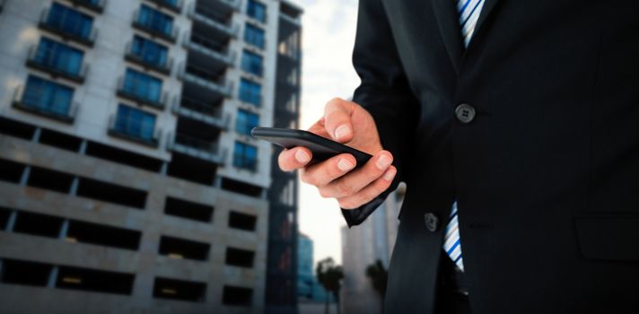 Midsection of businessman text messaging on mobile phone against glass modern building against cloudy sky