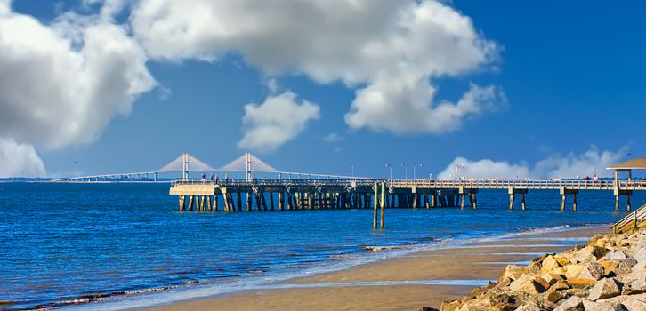 A white suspension bridge in distance beyond a wooden pier by a stone seawall on the beach with room for copy