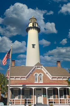 A white lighthouse againsta blue sky behind a brick home with an American Flag