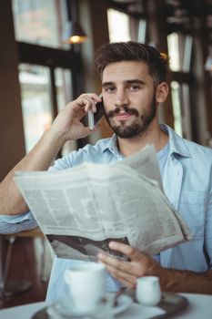 Portrait of confident executive talking on mobile phone while reading newspaper in cafÃ©