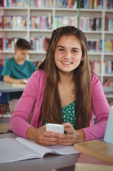 Portrait of smiling schoolgirl using mobile phone in library at school