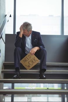 Depressed businessman sitting on stairs holding cardboard sheet with text need work at office