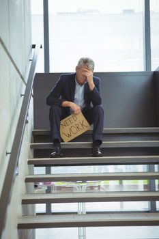 Depressed businessman sitting on stairs holding cardboard sheet with text need work at office