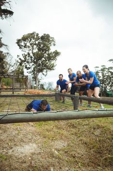Fit man crawling under the net during obstacle course while fit people cheering in bootcamp