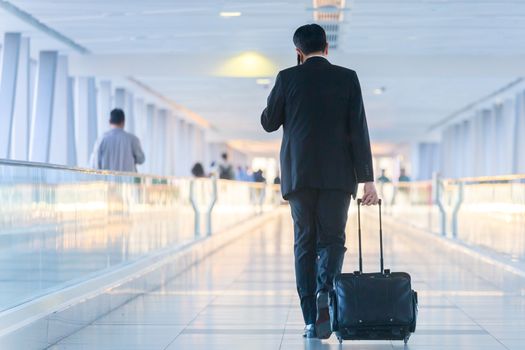 Rear view of unrecognizable formaly dressed businessman walking and wheeling a trolley suitcase at the lobby, talking on a mobile phone. Business travel concept.