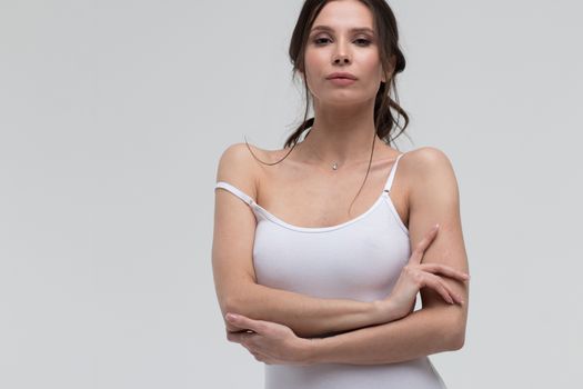 Relaxed sensual brunette woman in white t shirt pulling thin strap while looking at camera on white background