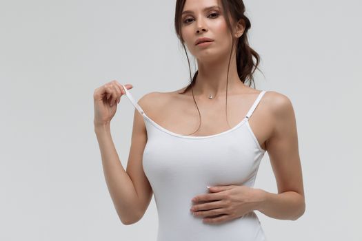Relaxed sensual brunette woman in white t shirt pulling thin strap while looking at camera on white background