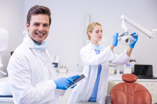 Dentist using digital tablet while his colleague adjusting dental light in background at clinic