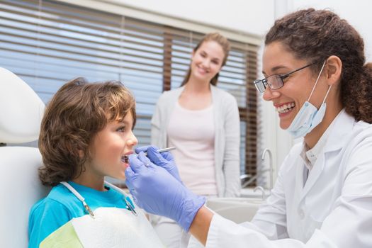 Pediatric dentist examining a little boys teeth with his mother watching at the dental clinic