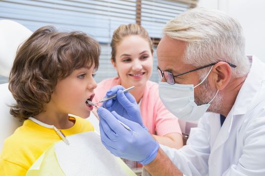 Pediatric dentist examining a little boys teeth with his mother watching at the dental clinic