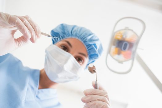 Dentist in surgical mask and cap holding dental tools over patient at the dental clinic
