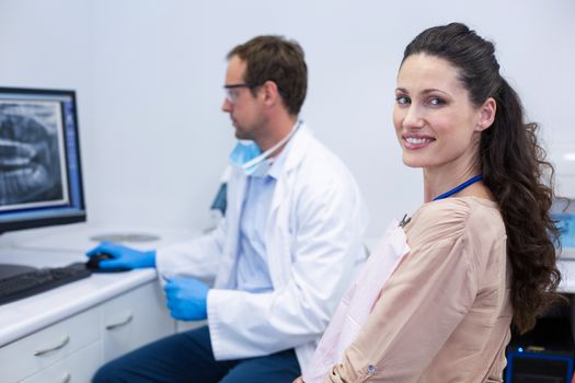Female patient smiling at camera while dentist looking at an x-ray at dental clinic
