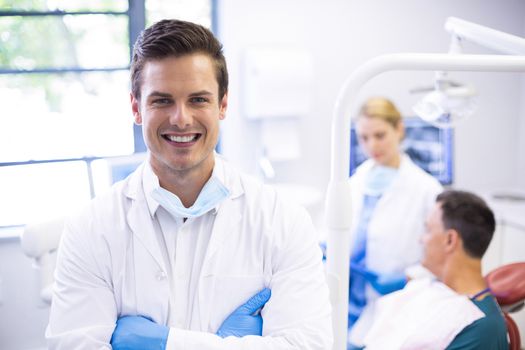 Portrait of dentist standing with arms crossed while his colleague examining patient in background