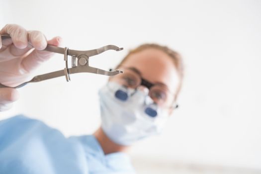 Dentist in surgical mask and dental loupes holding pliers over patient at the dental clinic