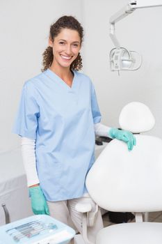 Dentist in blue scrubs smiling at camera beside chair at the dental clinic