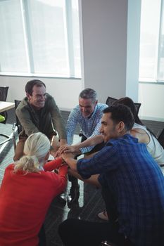 Happy business people stacking hands while sitting on chairs by window at office