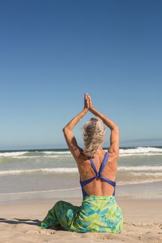 Rear view of senior woman with arms raised practising yoga while sitting on shore at beach