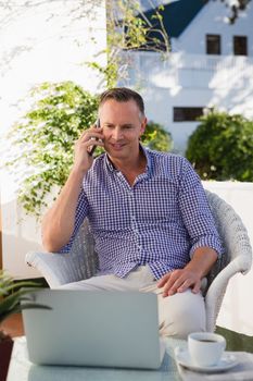 Mature businessman talking on mobile phone while using laptop in outdoors cafe