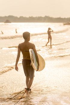 Silhouette of unrecognizable male surfer walking at tropical beach of Midigama, Sri Lankha at sunsen with surf board in his hands.