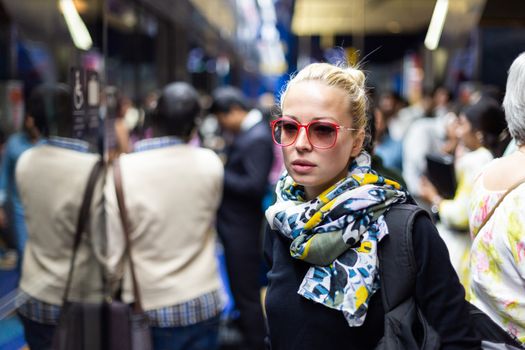 Young woman wearing colorful scarf waiting on the platform of a urban metro station for train to arrive. Public transport.