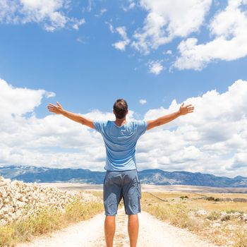 Rear view of casual sporty man standing on a dirt country road rising hands up to the clouds on a blue summer sky. Freedom and travel adventure concept. Copy space.