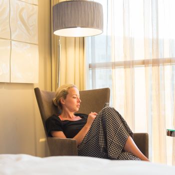 Businesswoman on business travel, tired from business meeting, sitting alone in hotel room afterwork, reading and responding to emails and messages on her smart phone.