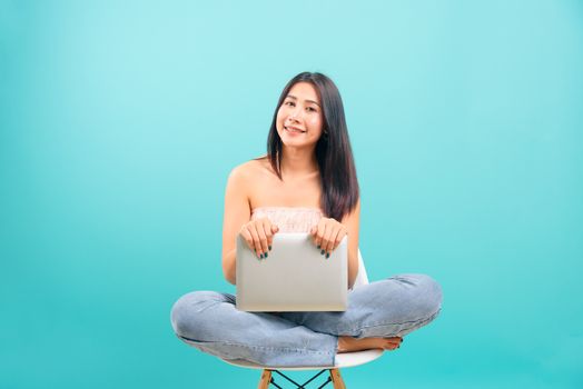 Smiling face portrait asian beautiful woman sitting on white chair with crossed legs her using laptop computer on blue background, with copy space for text