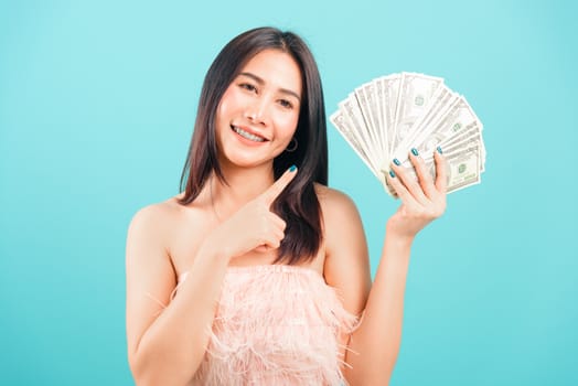 Smiling face portrait asian beautiful woman cheerful her holding and point the money on blue background, with copy space for text