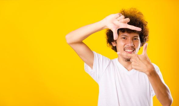 Asian handsome man with curly hair he smiling making frame with hands and fingers with happy face for creativity photography isolate on yellow background