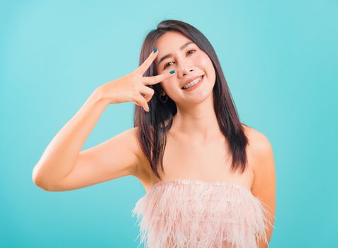 Portrait asian beautiful woman smiling showing peace gesture hand v-sign near eye good mood and her looking to camera on blue background