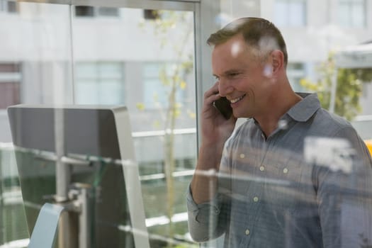 Smiling fashion designer talking on mobile phone by computer seen through glass