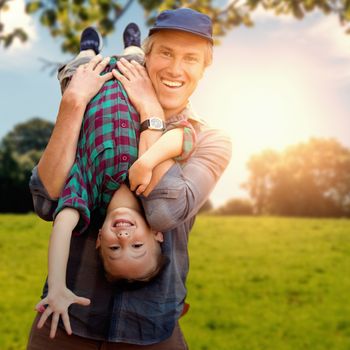 father holding his son upside down against green field against a green forest ans blue sky 