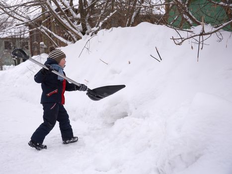 A boy in winter in warm clothes plays with a big shovel in the snow.