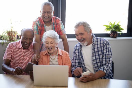 Happy senior people using technology while sitting at table against window in nursing home