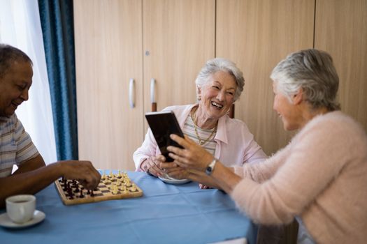 Smiling senior friends playing chess and using technology while sitting at table in nursing home