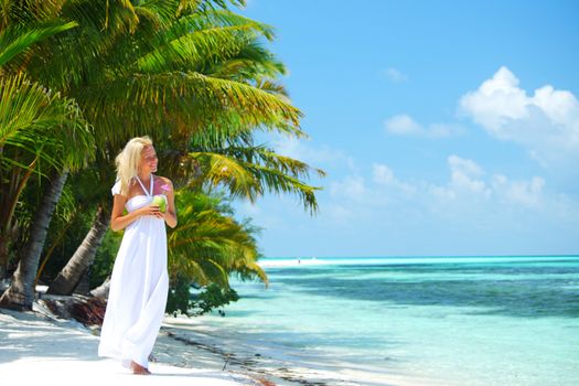 Woman in long white dress walking on tropical beach with palm trees holding coconut cocktail and smiling