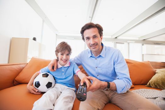 Cheerful father and son watching soccer match together at home holding ball