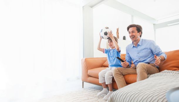 Cheerful father and son watching soccer match together at home holding ball