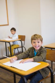 Pupil writing in notepad at his desk smiling at camera at the elementary school