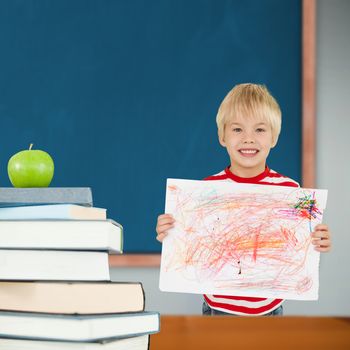 Cute boy showing his art against green apple on pile of books in classroom