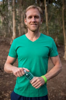 Portrait of smiling young man standing in the forest and holding water bottle in his hands