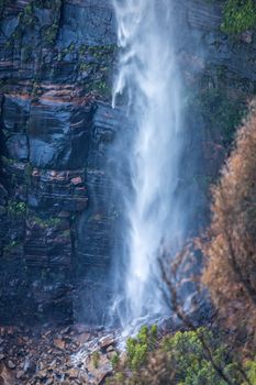 A waterfall tumbling over sheer cliff onto a rubble of rocks and boulders at its base in the Blue Mountains.  A short exposure the motion in vegetatation is from wind but has an arty feel