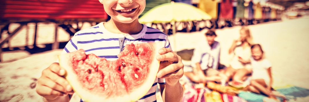 Portrait of smiling boy holding watermelon with family sitting in background at beach during sunny day