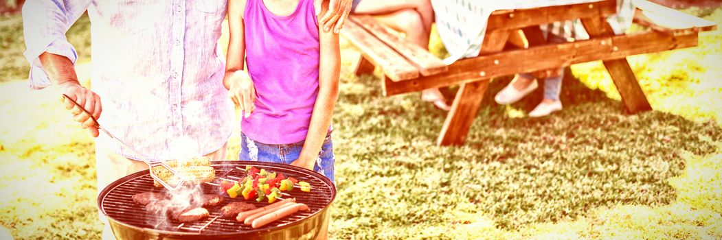 Grandfather and granddaughter preparing barbecue while family having meal in background