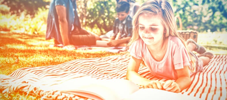 Girl lying on blanket and reading book while family sitting in background on a sunny day